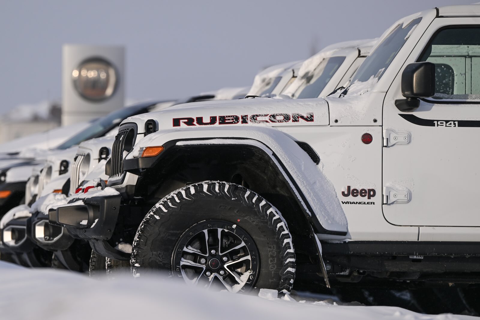 White Jeep Wranglers lined up in the snow at a dealership, a sign visible in the background.