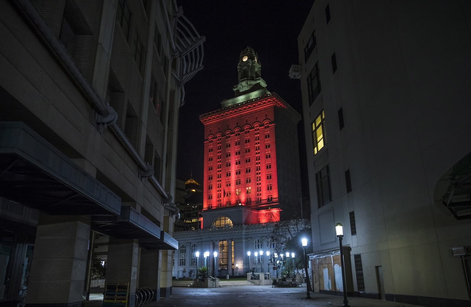 Red lights illuminate the Oakland City Hall, the buildings of a narrow alleyway visible in the foreground.