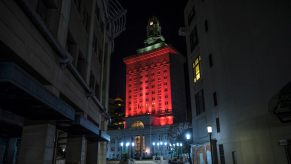 Red lights illuminate the Oakland City Hall, the buildings of a narrow alleyway visible in the foreground.