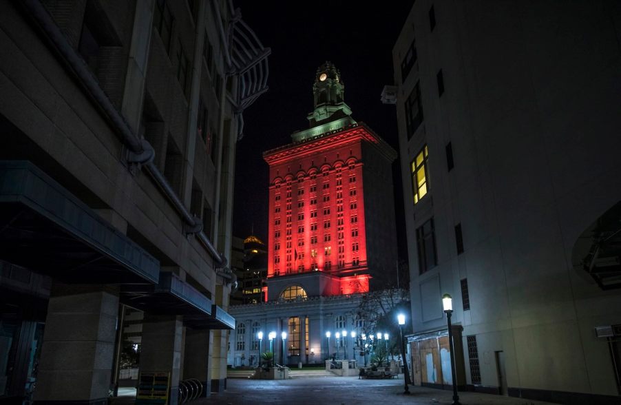 Red lights illuminate the Oakland City Hall, the buildings of a narrow alleyway visible in the foreground.