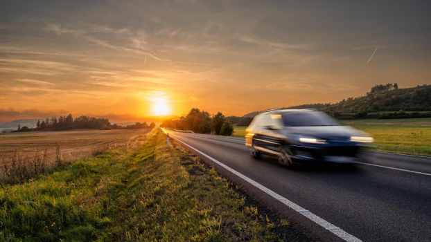 Sun sets behind a speeding car that's a blur on a rural highway.