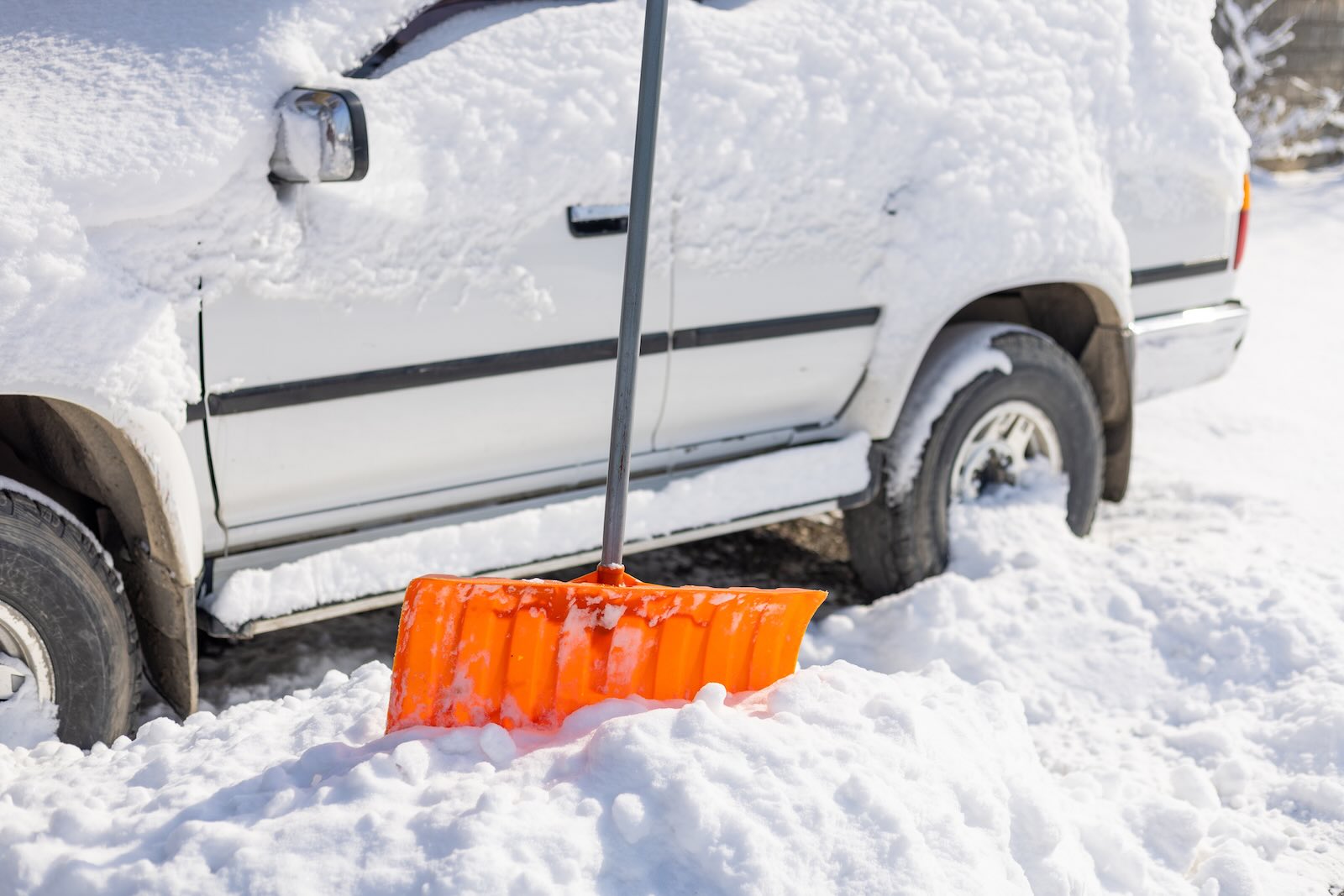 Orange shovel in front of white SUV covered in snow.
