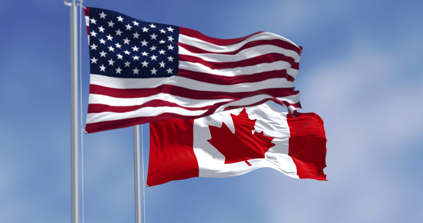 National flags of U.S. and Canada flying next to one another, blue sky in the background.