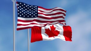 National flags of U.S. and Canada flying next to one another, blue sky in the background.