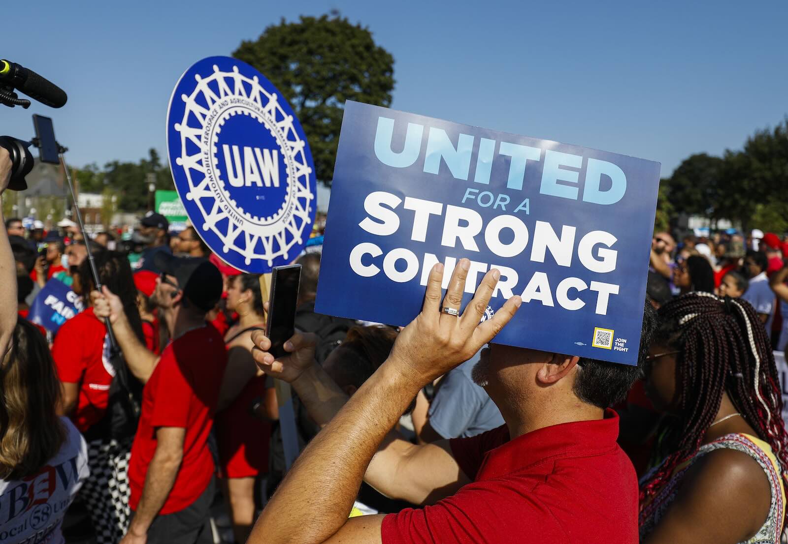 UAW members hold up signs during a union strike, the blue sky visible in the background.