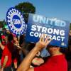 UAW members hold up signs during a union strike, the blue sky visible in the background.