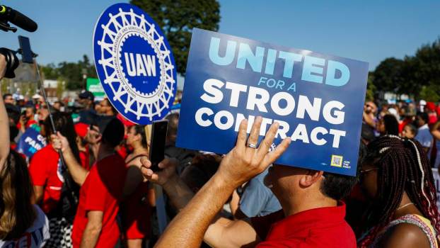 UAW members hold up signs during a union strike, the blue sky visible in the background.