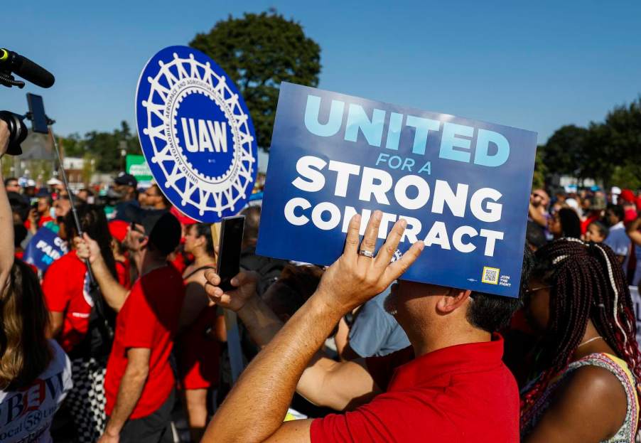 UAW members hold up signs during a union strike, the blue sky visible in the background.