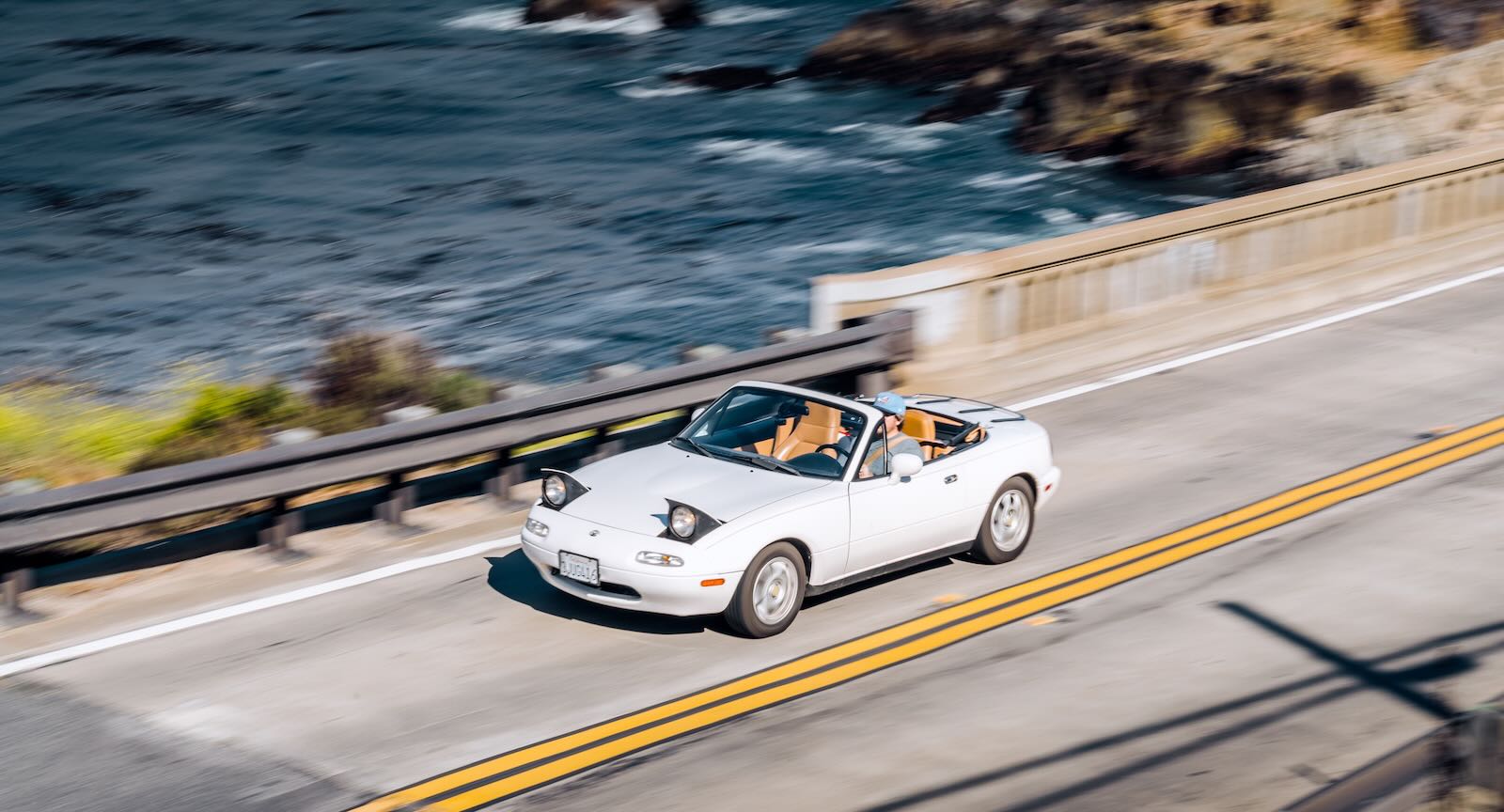 White Mazda Miata roadster convertible on a bridge, the Pacific Ocean visible in the background.