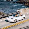 White Mazda Miata roadster convertible on a bridge, the Pacific Ocean visible in the background.
