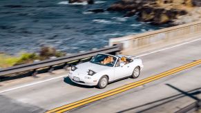 White Mazda Miata roadster convertible on a bridge, the Pacific Ocean visible in the background.