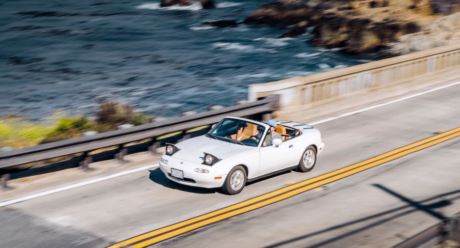 White Mazda Miata roadster convertible on a bridge, the Pacific Ocean visible in the background.