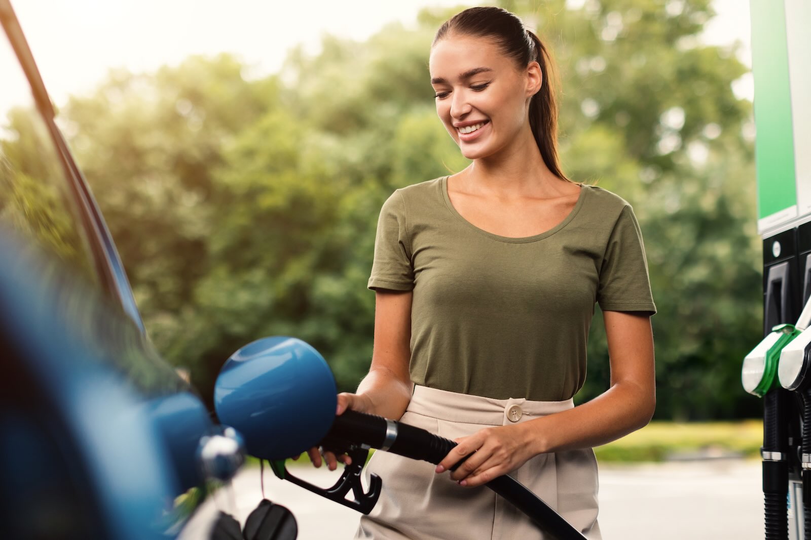 Woman fills up her fuel tank at a gas station, trees visible in the background.