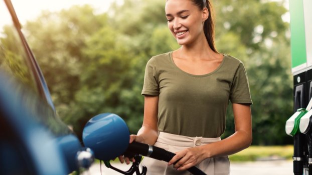 Woman fills up her fuel tank at a gas station, trees visible in the background.