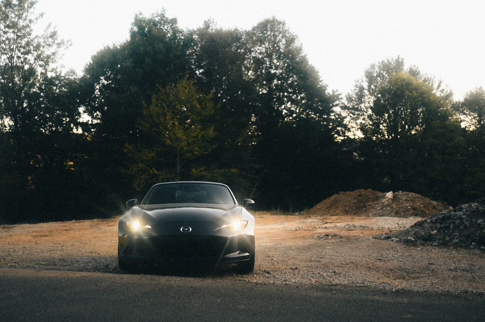 A black car with headlights on parked outdoors at twilight surrounded by trees.